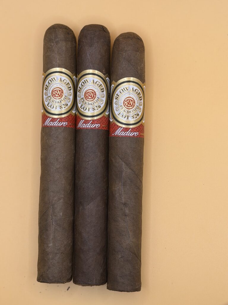 Three dark brown cigars with gold-embossed bands and a red Maduro ribbon, lined up vertically against a peach background.