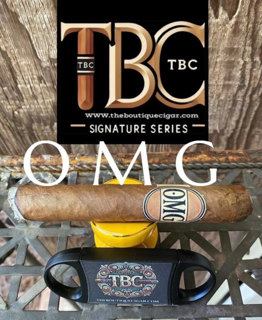 Cigar resting on a yellow stand with a black promo sign behind, large white OMG text across the foreground, branding for The Boutique Cigar (TBC).
