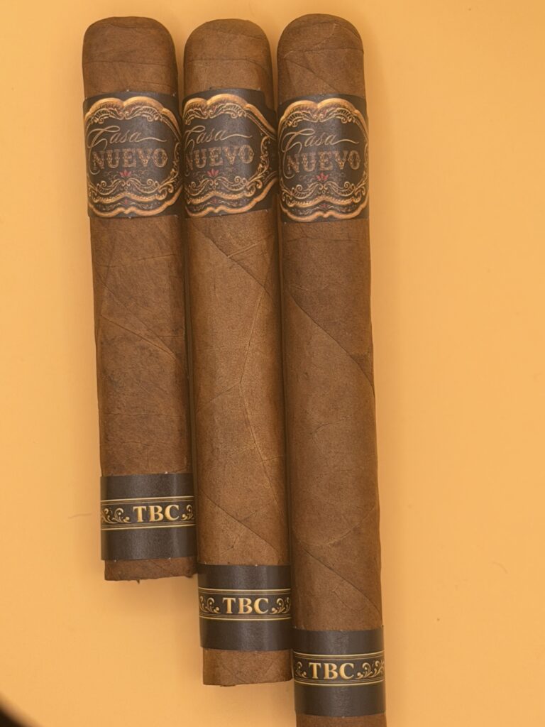 Three cigars with ornate black-and-gold bands reading 'Casa Nuevo' and 'TBC' stand side by side on an orange background.